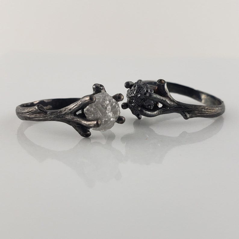 Puede incluir: Dos anillos de plata con un dise&ntilde;o similar a una ramita. Un anillo presenta una piedra clara y en bruto, mientras que el otro tiene una piedra oscura y texturizada. Los anillos se colocan sobre un fondo blanco, mostrando su est&eacute;tica &uacute;nica inspirada en la naturaleza.