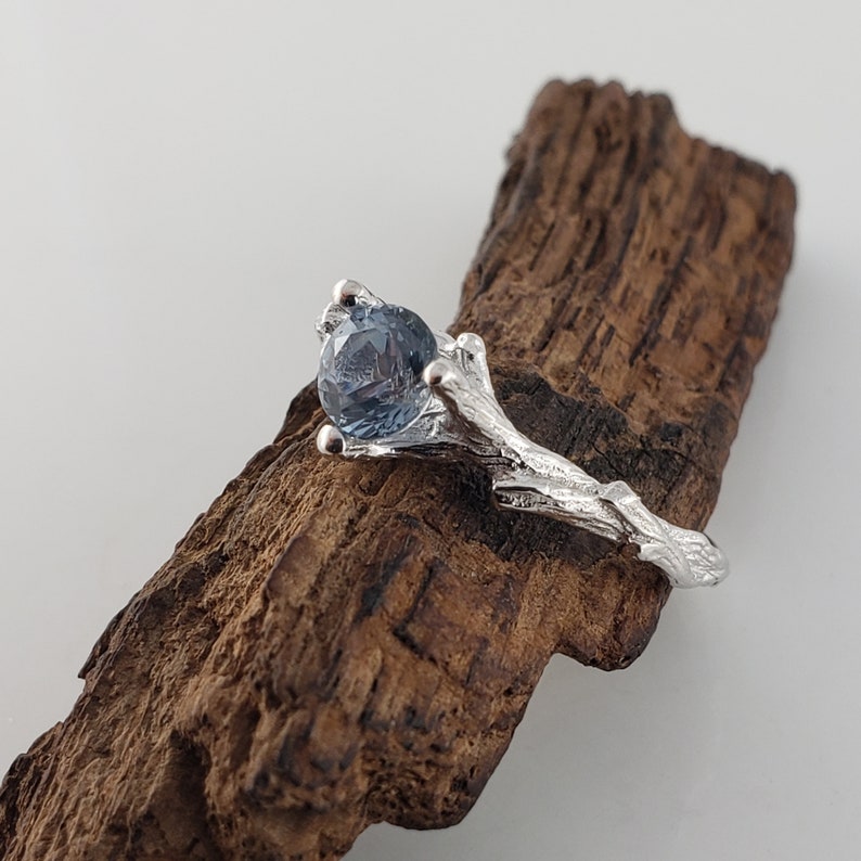May include: A silver ring featuring a blue gemstone set in a branch-like design. The ring is displayed on a piece of dark wood.