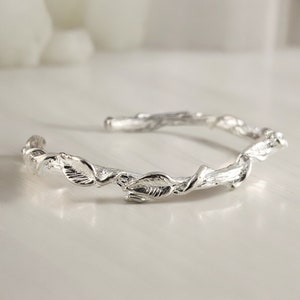 May include: A silver bangle bracelet with a twisted branch design and leaves. The bracelet is open and has a delicate, nature-inspired look.