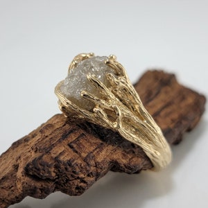 May include: A gold ring featuring a rough-cut, light gray gemstone. The ring's band and setting have a textured, organic design, resembling intertwined branches. The ring is displayed on a piece of wood.