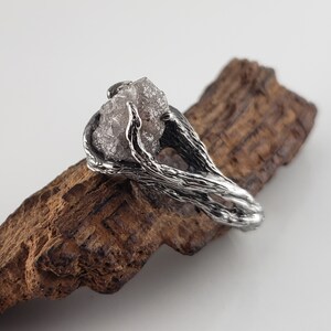 Op de afbeelding: Een zilveren ring met een ruwe, grijze edelsteen, gehouden door een getextureerde, takachtige zetting. De ring wordt gepresenteerd op een stuk donkerbruin hout tegen een witte achtergrond. Het ontwerp heeft een natuurlijke, organische uitstraling.