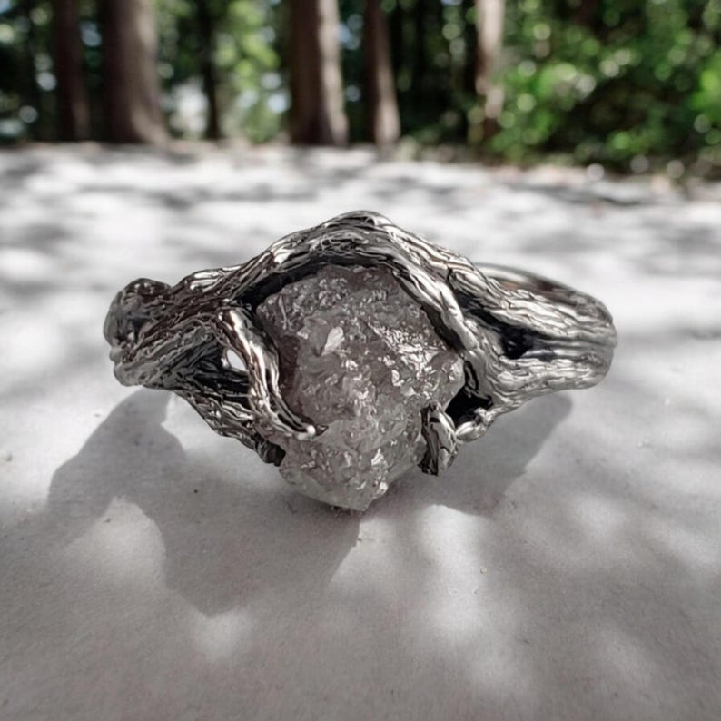 Op de afbeelding: Een zilverkleurige ring met een ruw geslepen, lichtgekleurde edelsteen. De band van de ring is ontworpen om verweven takken na te bootsen, wat een natuurlijke, organische uitstraling cre&euml;ert. De edelsteen staat centraal.