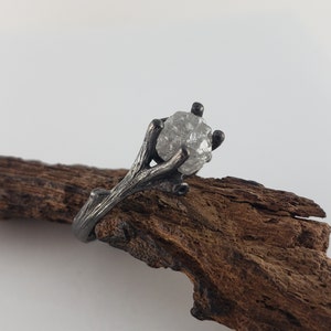 Puede incluir: Un anillo plateado con una gema en bruto y transparente. La banda y el engaste del anillo tienen un dise&ntilde;o similar a una rama, con la piedra sujeta por garras. El anillo se muestra sobre un trozo de madera marr&oacute;n oscuro.