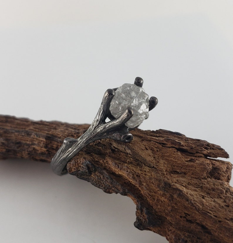 Puede incluir: Un anillo plateado con una gema en bruto y transparente. La banda y el engaste del anillo tienen un dise&ntilde;o similar a una rama, con la piedra sujeta por garras. El anillo se muestra sobre un trozo de madera marr&oacute;n oscuro.