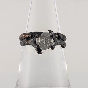 Puede incluir: Un anillo plateado con una gema en bruto sin cortar en el centro. La banda y el engaste del anillo tienen un dise&ntilde;o texturizado en forma de rama, con detalles oscuros. La gema es de color gris claro y est&aacute; sujeta por peque&ntilde;as garras oscuras.