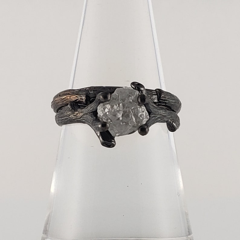 Puede incluir: Un anillo plateado con una gema en bruto sin cortar en el centro. La banda y el engaste del anillo tienen un dise&ntilde;o texturizado en forma de rama, con detalles oscuros. La gema es de color gris claro y est&aacute; sujeta por peque&ntilde;as garras oscuras.