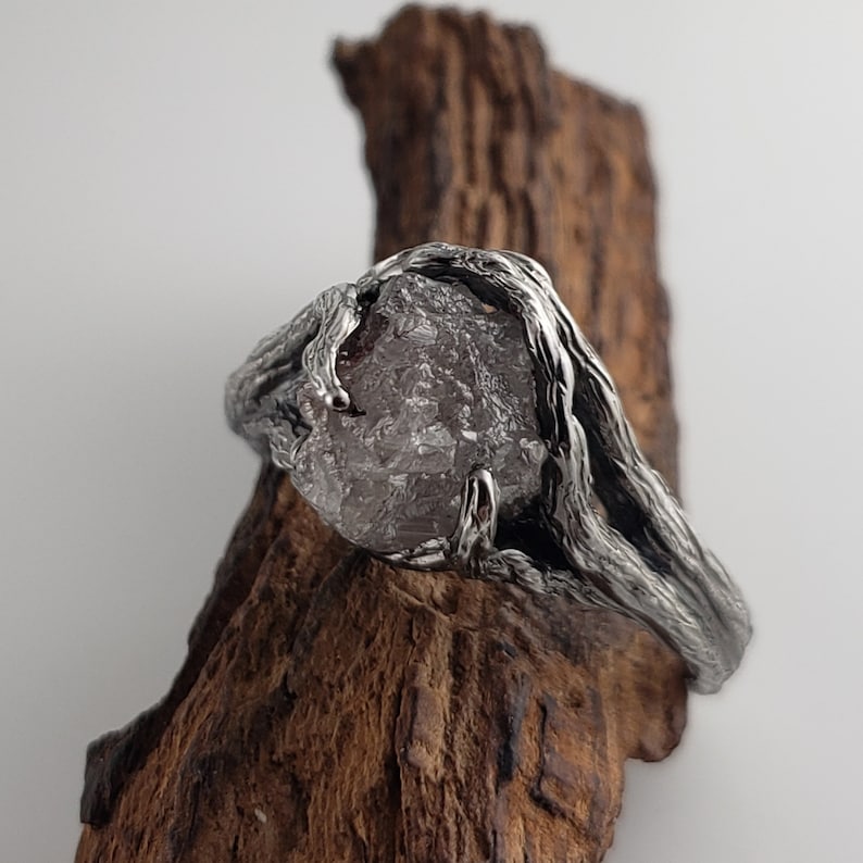 Op de afbeelding: Een zilveren ring met een grote, ruw geslepen, grijze edelsteen. De band van de ring heeft een textuur, organisch ontwerp dat om de steen gewikkeld is. De ring wordt getoond op een stuk donkerbruin hout.