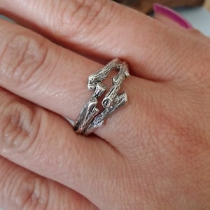May include: A silver ring featuring a design of interwoven branches. The ring is worn on a person's finger.