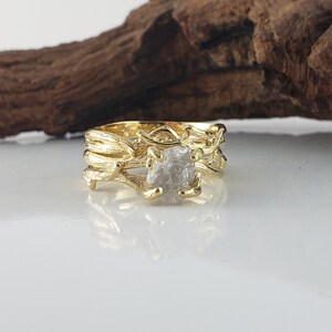 May include: A gold ring with a rough, white diamond set in a branch-like design. The ring is made of two bands, one with a thicker branch design and the other with a thinner branch design.