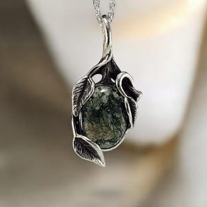 May include: A silver pendant necklace featuring an oval moss agate gemstone with dark green inclusions. The stone is encased in a detailed silver setting shaped like twisted branches and leaves, suspended from a delicate silver chain link necklace.
