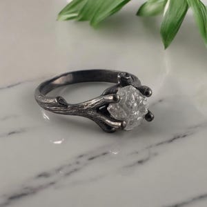 Puede incluir: Un anillo plateado con una gema clara y en bruto. El dise&ntilde;o del anillo imita una ramita, con la banda y el engaste formados como ramas que sostienen la piedra. El anillo se muestra sobre una superficie de m&aacute;rmol blanco.