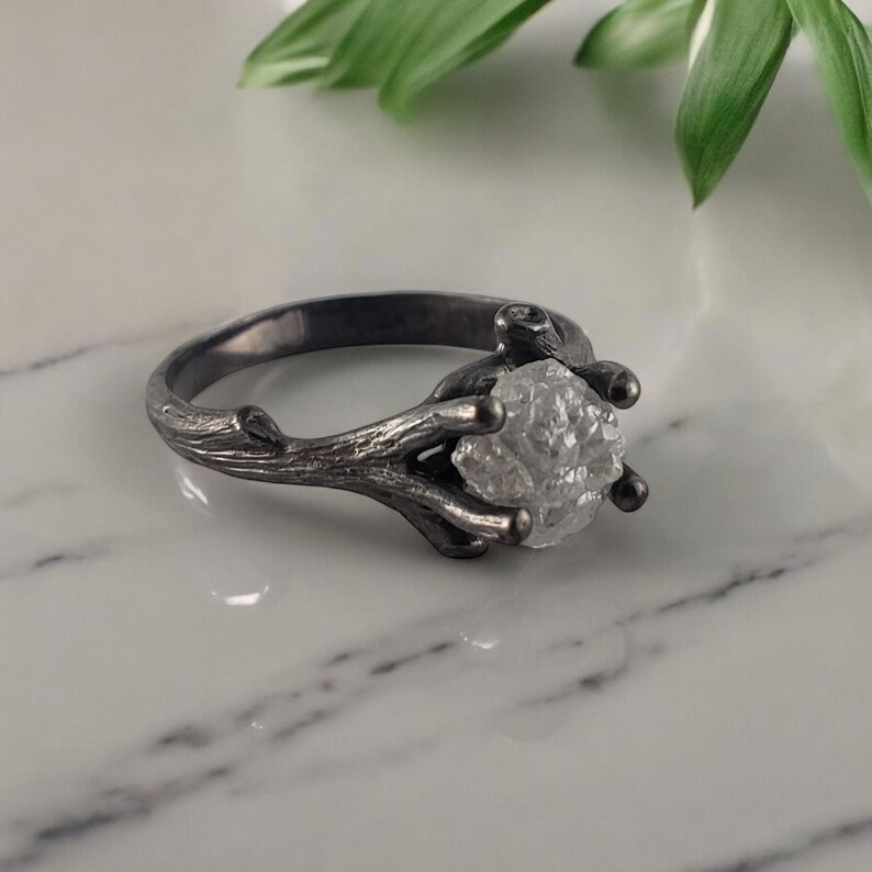 Puede incluir: Un anillo plateado con una gema clara y en bruto. El dise&ntilde;o del anillo imita una ramita, con la banda y el engaste formados como ramas que sostienen la piedra. El anillo se muestra sobre una superficie de m&aacute;rmol blanco.