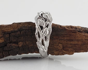 Puede incluir: Un anillo de plata con un delicado diseño de hojas y ramas. El anillo se muestra sobre un trozo de madera flotante.