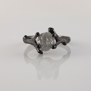 Puede incluir: Un anillo de plata con una gema en bruto de color gris claro. La banda del anillo tiene un dise&ntilde;o texturizado en forma de rama con detalles oscuros. La gema est&aacute; sujeta por las ramas, creando una joya &uacute;nica inspirada en la naturaleza.