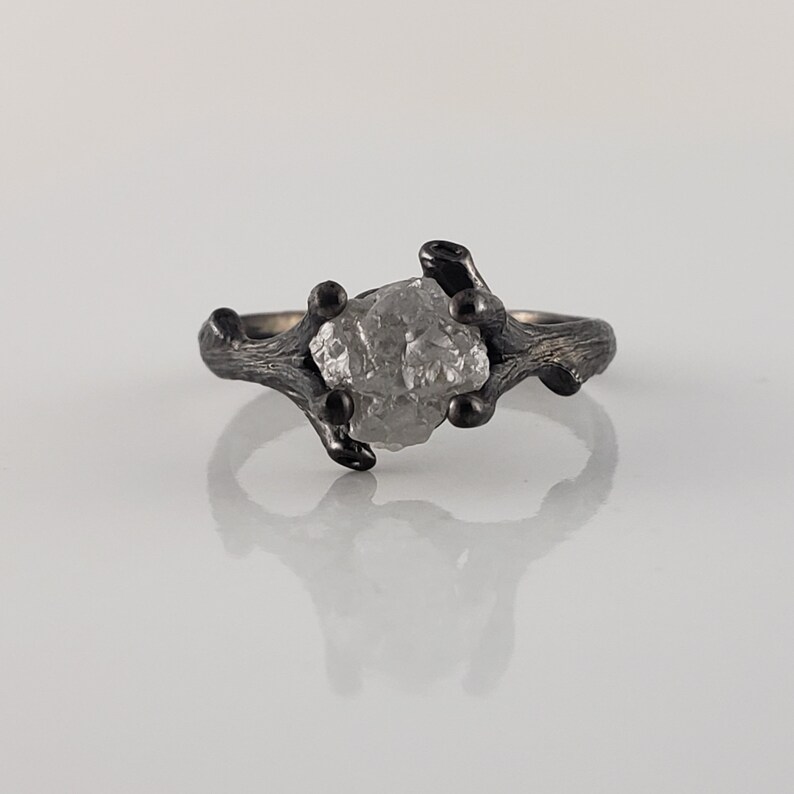 Puede incluir: Un anillo de plata con una gema en bruto de color gris claro. La banda del anillo tiene un dise&ntilde;o texturizado en forma de rama con detalles oscuros. La gema est&aacute; sujeta por las ramas, creando una joya &uacute;nica inspirada en la naturaleza.