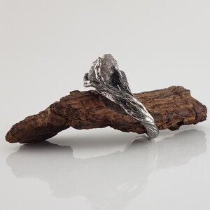 Op de afbeelding: Een zilveren ring met een ruw, natuurlijk ogend ontwerp. De ring heeft een grote, onregelmatig gevormde, lichtgekleurde steen, gezet in een getextureerde band die lijkt op verweven takken. De ring wordt getoond op een stuk hout.