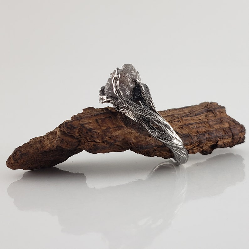 Op de afbeelding: Een zilveren ring met een ruw, natuurlijk ogend ontwerp. De ring heeft een grote, onregelmatig gevormde, lichtgekleurde steen, gezet in een getextureerde band die lijkt op verweven takken. De ring wordt getoond op een stuk hout.