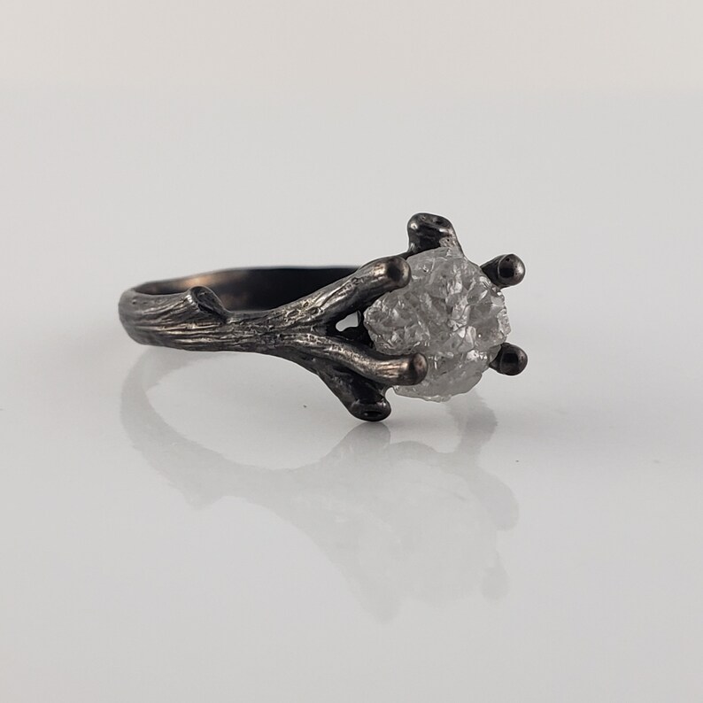 Puede incluir: Un anillo plateado con una gema en bruto de color gris claro. La banda y el engaste del anillo est&aacute;n dise&ntilde;ados para parecerse a ramas de &aacute;rbol, creando una est&eacute;tica inspirada en la naturaleza. La gema est&aacute; sujeta por seis garras.