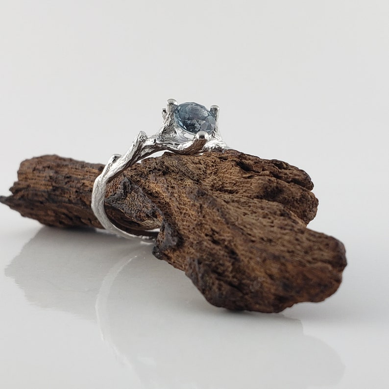 May include: A silver ring featuring a blue gemstone set in a branch-like design. The ring is resting on a piece of dark brown driftwood.