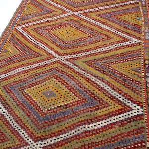 May include: A rectangular woven rug featuring a repeating geometric pattern of large diamonds. The design incorporates a vibrant color palette of mustard yellow, deep red, olive green, and purple, accented by thin white lines and small dotted details.