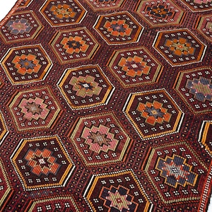May include: A rectangular woven rug featuring a repeating pattern of dark red hexagons. Each hexagon contains a central geometric star motif in shades of orange, yellow, blue, and pink, outlined by thin white and yellow borders against a dark background.
