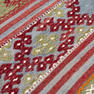 May include: Close-up of a woven rug with a geometric pattern. The rug features a combination of red, blue, and green hues. Diagonal red stripes and diamond shapes create a visually appealing design. The rug is likely handmade.
