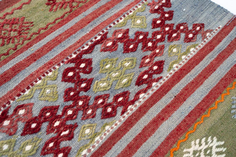 May include: Close-up of a woven rug with a geometric pattern. The rug features a combination of red, blue, and green hues. Diagonal red stripes and diamond shapes create a visually appealing design. The rug is likely handmade.