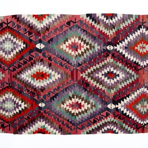 Puede incluir: Una alfombra rectangular tejida a mano con un patr&oacute;n geom&eacute;trico de diamantes. La alfombra presenta una vibrante paleta de colores rojo, morado, verde y blanco. El dise&ntilde;o incluye formas de diamantes repetidas y bordes en zigzag.