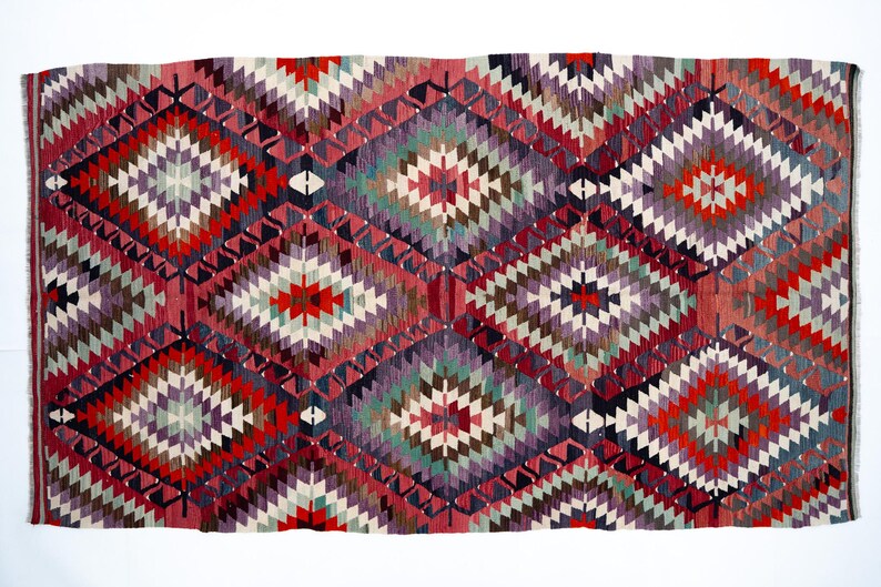 Puede incluir: Una alfombra rectangular tejida a mano con un patr&oacute;n geom&eacute;trico de diamantes. La alfombra presenta una vibrante paleta de colores rojo, morado, verde y blanco. El dise&ntilde;o incluye formas de diamantes repetidas y bordes en zigzag.