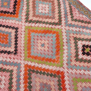 May include: A close-up of a colorful, geometric patterned rug. The rug features a repeating square design with pink, orange, green, and blue hues. The edges are finished with a decorative border.