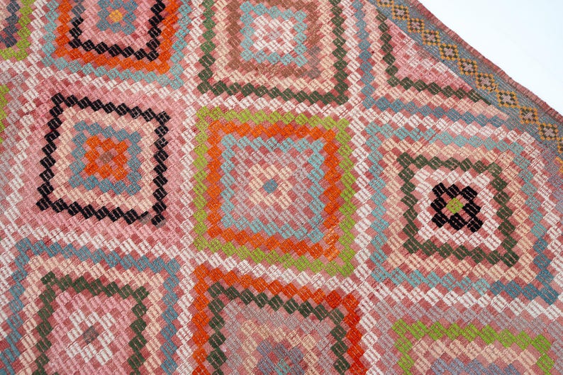 May include: A close-up of a colorful, geometric patterned rug. The rug features a repeating square design with pink, orange, green, and blue hues. The edges are finished with a decorative border.