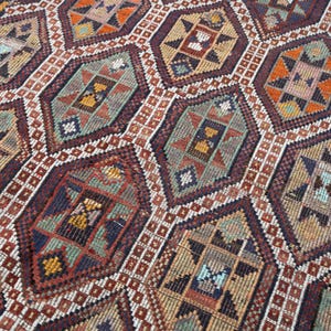 May include: A detailed close-up of a woven rug with a geometric pattern. The rug features a repeating design of diamond and hexagonal shapes in shades of brown, red, orange, and blue. The intricate pattern suggests a traditional or vintage style.