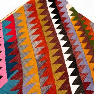 K&ouml;nnte beinhalten: Ein farbenfrohes, geometrisches Textil mit einem sich wiederholenden Zickzackmuster in verschiedenen Farben. Das Design umfasst rote, orange, gelbe, gr&uuml;ne, blaue und schwarze Dreiecke. Das Textil scheint gewebt zu sein.
