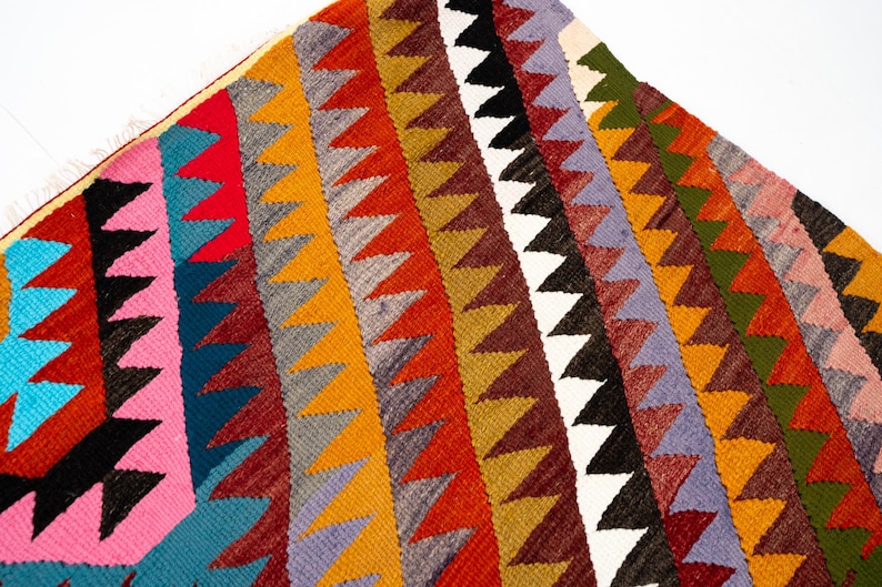 K&ouml;nnte beinhalten: Ein farbenfrohes, geometrisches Textil mit einem sich wiederholenden Zickzackmuster in verschiedenen Farben. Das Design umfasst rote, orange, gelbe, gr&uuml;ne, blaue und schwarze Dreiecke. Das Textil scheint gewebt zu sein.