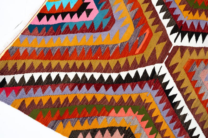 K&ouml;nnte beinhalten: Ein farbenfroher, geometrisch gemusterter Teppich mit einer Vielzahl von Farben, darunter Orange, Rot, Gelb, Blau, Rosa und Schwarz. Der Teppich weist ein sich wiederholendes Zickzack-Muster auf, das eine strukturierte Oberfl&auml;che erzeugt. Der Teppich ist wahrscheinlich aus gewebtem Stoff.