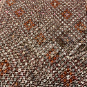 May include: A woven textile rug featuring a dense, repeating geometric grid pattern in shades of dark brown, orange, white, and grey. The edges are finished with a decorative border containing intricate pink, white, and orange diamond motifs.
