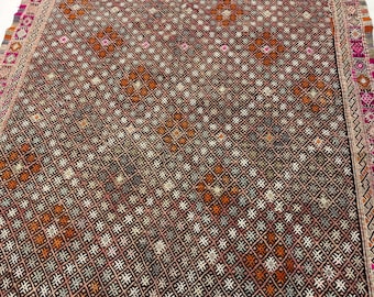 May include: A woven textile rug featuring a dense, repeating geometric grid pattern in shades of dark brown, orange, white, and grey. The edges are finished with a decorative border containing intricate pink, white, and orange diamond motifs.