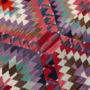 Puede incluir: Primer plano de una alfombra con estampado geom&eacute;trico colorido. La alfombra presenta un patr&oacute;n de diamantes repetido en tonos de rojo, morado, verde azulado, marr&oacute;n y blanco. El dise&ntilde;o es intrincado y detallado, con una apariencia texturizada.