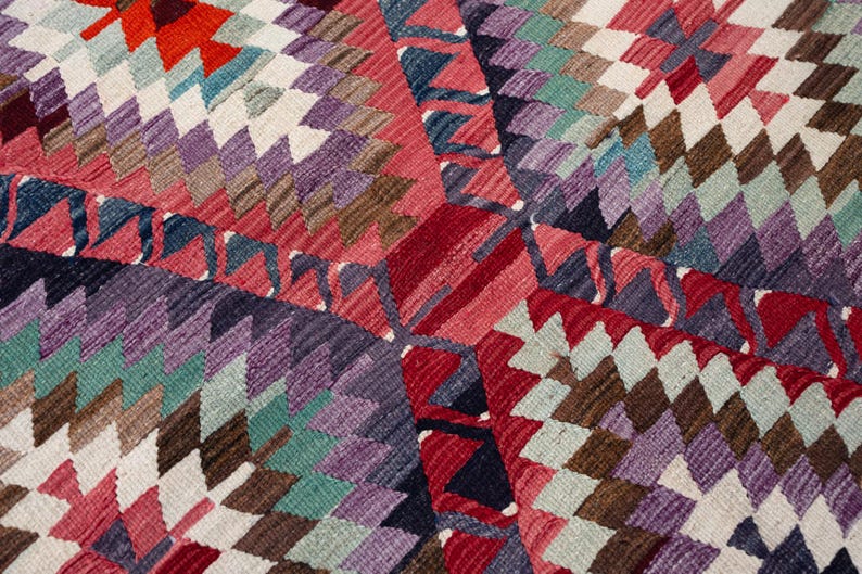 Puede incluir: Primer plano de una alfombra con estampado geom&eacute;trico colorido. La alfombra presenta un patr&oacute;n de diamantes repetido en tonos de rojo, morado, verde azulado, marr&oacute;n y blanco. El dise&ntilde;o es intrincado y detallado, con una apariencia texturizada.