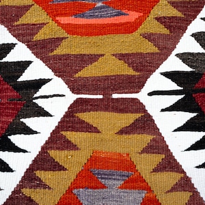 K&ouml;nnte beinhalten: Nahaufnahme eines gewebten Textils mit geometrischem Muster. Das Design zeigt sich wiederholende Rautenformen mit Dreiecken in Rot, Wei&szlig;, Schwarz, Gold und Lila. Das Textil hat ein strukturiertes Aussehen.