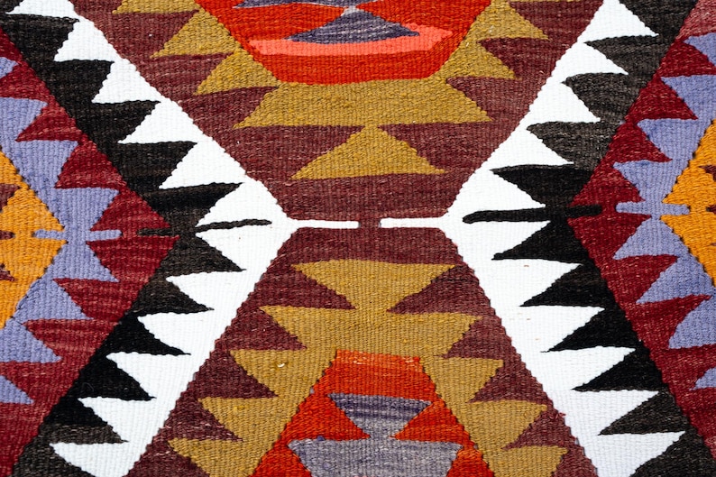 K&ouml;nnte beinhalten: Nahaufnahme eines gewebten Textils mit geometrischem Muster. Das Design zeigt sich wiederholende Rautenformen mit Dreiecken in Rot, Wei&szlig;, Schwarz, Gold und Lila. Das Textil hat ein strukturiertes Aussehen.