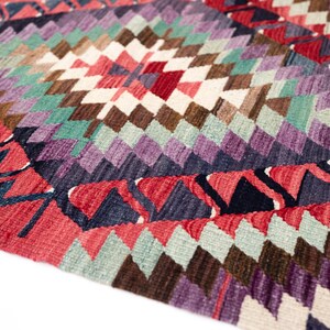 Puede incluir: Primer plano de una colorida alfombra tejida con un patr&oacute;n geom&eacute;trico de diamantes. La alfombra presenta una variedad de colores, incluyendo rojo, morado, verde, marr&oacute;n y blanco. Los bordes tienen un dise&ntilde;o en zigzag. Esta alfombra es perfecta para a&ntilde;adir un toque de color a cualquier habitaci&oacute;n.