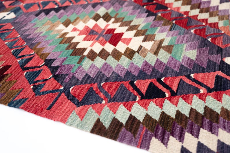 Puede incluir: Primer plano de una colorida alfombra tejida con un patr&oacute;n geom&eacute;trico de diamantes. La alfombra presenta una variedad de colores, incluyendo rojo, morado, verde, marr&oacute;n y blanco. Los bordes tienen un dise&ntilde;o en zigzag. Esta alfombra es perfecta para a&ntilde;adir un toque de color a cualquier habitaci&oacute;n.
