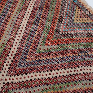 May include: Close-up of a colourful, patterned rug with a geometric design. The rug features a mix of red, green, blue, and white hues, creating a vibrant and textured surface. The edges have a fringed border.