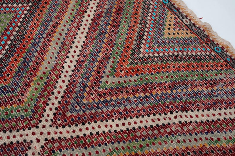 May include: Close-up of a colourful, patterned rug with a geometric design. The rug features a mix of red, green, blue, and white hues, creating a vibrant and textured surface. The edges have a fringed border.