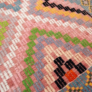 May include: Close-up of a woven rug with a geometric pattern. The rug features a mix of colors, including pink, green, white, blue, orange, and black. The design includes diamond and zigzag shapes, creating a textured surface.