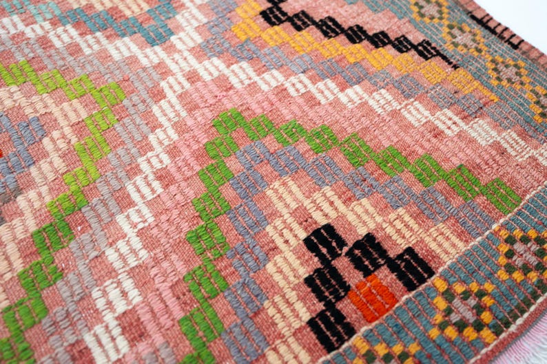 May include: Close-up of a woven rug with a geometric pattern. The rug features a mix of colors, including pink, green, white, blue, orange, and black. The design includes diamond and zigzag shapes, creating a textured surface.