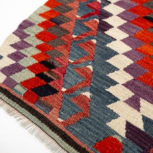 Puede incluir: Primer plano de una colorida alfombra tejida con un patr&oacute;n geom&eacute;trico de diamantes. La alfombra presenta una variedad de colores, incluyendo rojo, morado, azul y blanco. La alfombra tiene un borde con flecos y probablemente est&aacute; hecha de lana o un material similar.