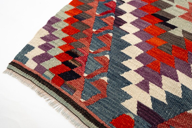 Puede incluir: Primer plano de una colorida alfombra tejida con un patr&oacute;n geom&eacute;trico de diamantes. La alfombra presenta una variedad de colores, incluyendo rojo, morado, azul y blanco. La alfombra tiene un borde con flecos y probablemente est&aacute; hecha de lana o un material similar.