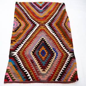 K&ouml;nnte beinhalten: Ein farbenfroher, geometrischer Teppich mit einem Rautenmuster. Der Teppich weist eine Vielzahl von Farben auf, darunter Rot, Blau, Gelb und Braun. Das Design umfasst Zickzack- und Dreiecksformen. Der Teppich ist rechteckig.